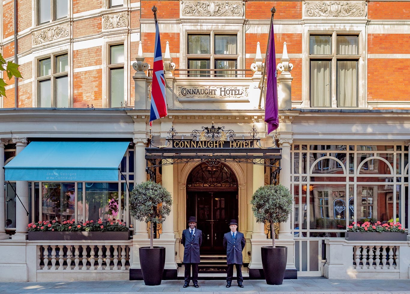 "The grand entrance of The Connaught Hotel in London, featuring elegant architecture, a classic black and gold canopy, two uniformed doormen, and the Union Jack and hotel flags displayed above.