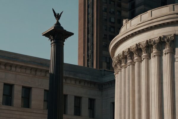 A neoclassical courthouse with grand columns and an ornate eagle-topped pillar, set against a backdrop of modern and historic buildings under a clear blue sky.