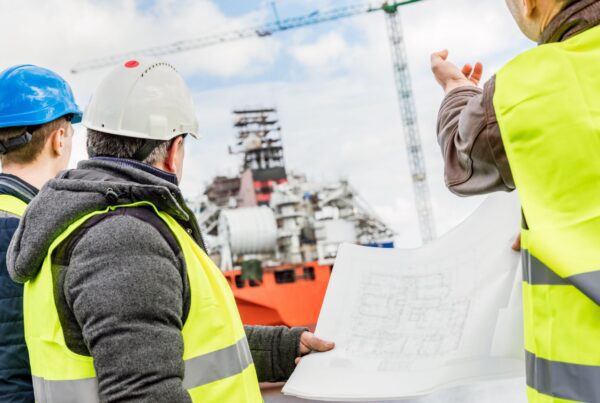 three shipbuilders looking at blueprints with ship in the background