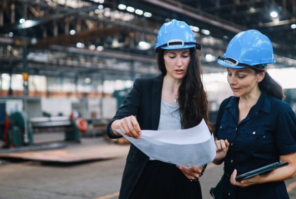 Two female engineers wearing blue hard hats examine blueprints in a factory setting.